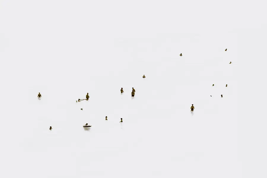 Photographie high-key en plan large : silhouettes de nageurs espacées dans l’eau calme, quelques reflets et une petite embarcation, composition minimaliste qui insiste sur le vide et la fragmentation des corps dans l’élément marin.