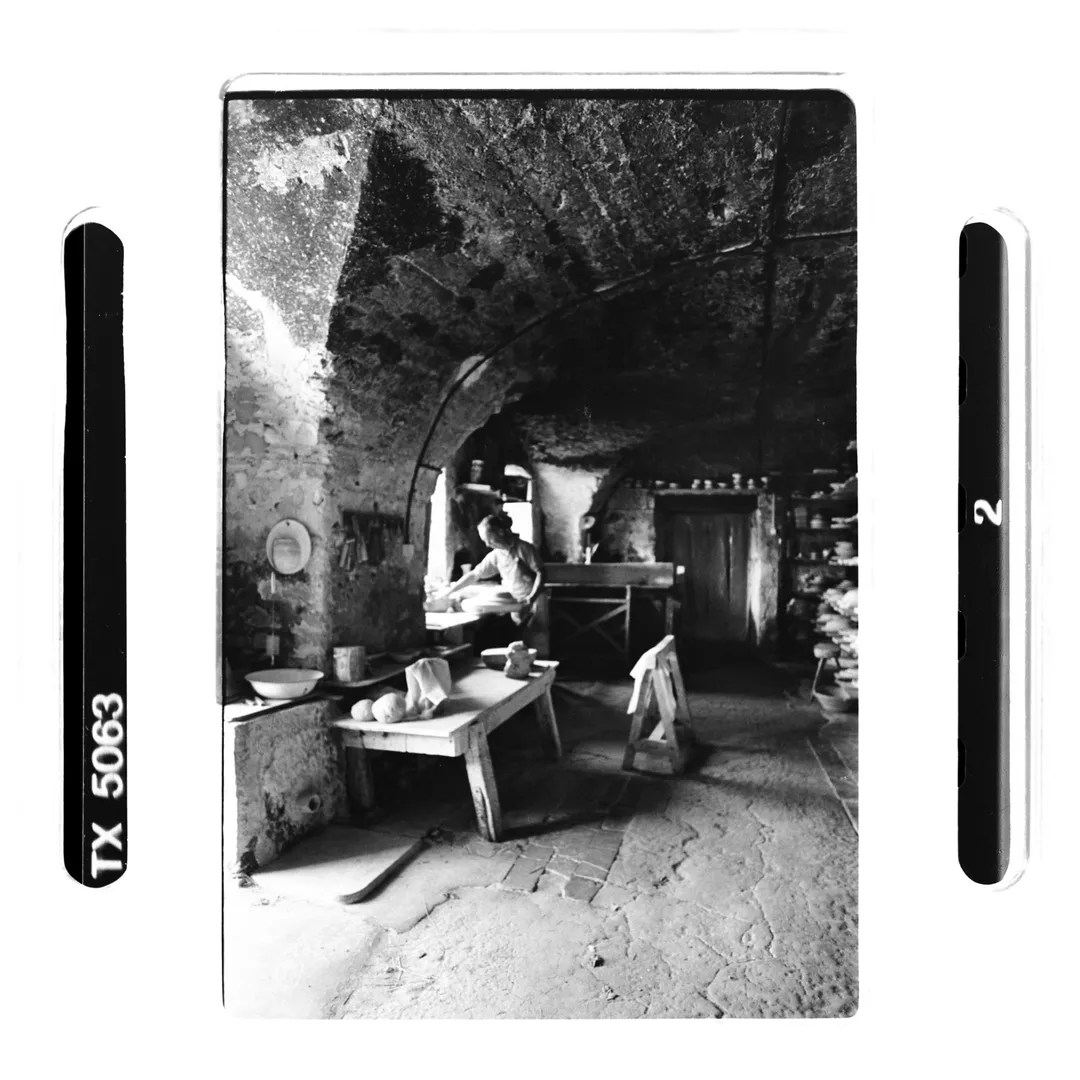 Photographie noir et blanc d’un atelier de poterie voûté au poët-laval (Drôme) avec un potier travaillant près de la fenêtre, établi en bois et étagères de céramiques, prise pour un reportage sur la poterie traditionnelle du village.