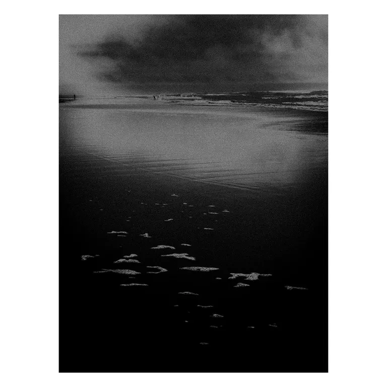 Photographie noir et blanc très contrastée d’une plage atlantique : plaques d’écume au premier plan sur le sable sombre, miroir d’eau calme vers l’horizon, vagues et nuages orageux en arrière-plan, série « Marée intérieure » de Nicolas Louis.