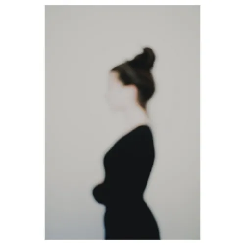 Photographie artistique défocalisée : silhouette féminine de profil, chignon et tenue sombre, contours adoucis et identité estompée, fond neutre minimaliste — extrait de la série « Erasure »