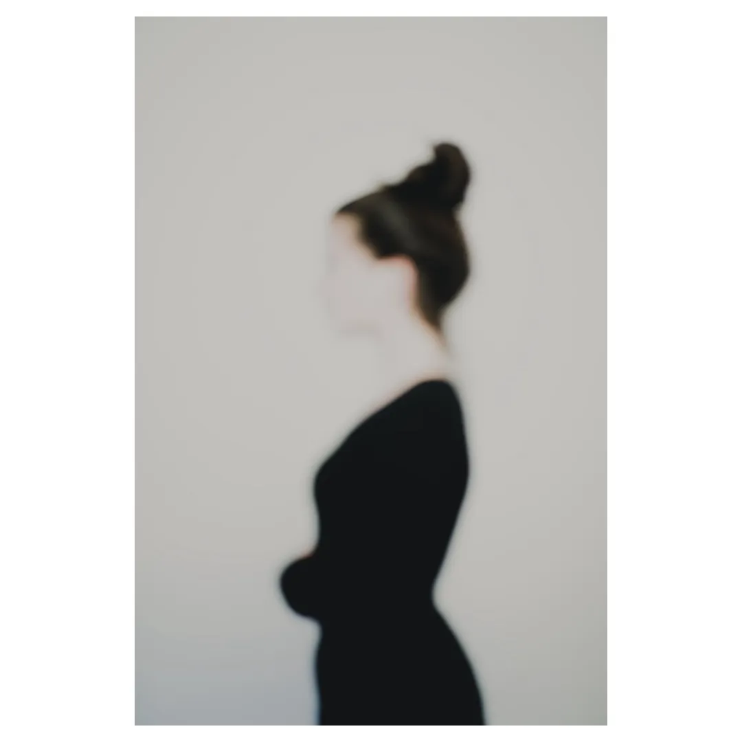 Photographie artistique défocalisée : silhouette féminine de profil, chignon et tenue sombre, contours adoucis et identité estompée, fond neutre minimaliste — extrait de la série « Erasure »
