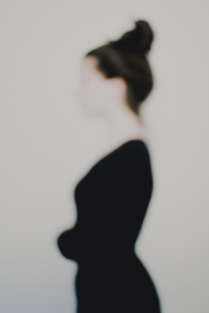 Photographie artistique défocalisée : silhouette féminine de profil, chignon et tenue sombre, contours adoucis et identité estompée, fond neutre minimaliste — extrait de la série « Erasure »