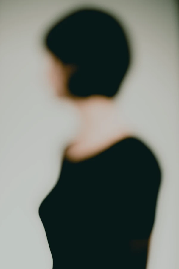 Portrait artistique défocalisé : femme aux cheveux courts vue de trois-quarts, buste sombre sur fond clair, contours adoucis et identité volontairement effacée — série « Erasure ».