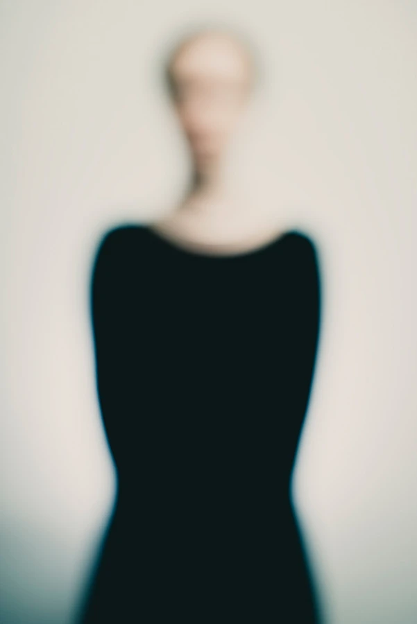Portrait artistique défocalisé : figure féminine de face, cou étiré et buste en robe noire, contours dissous sur fond neutre, identité volontairement estompée — série « Erasure ».