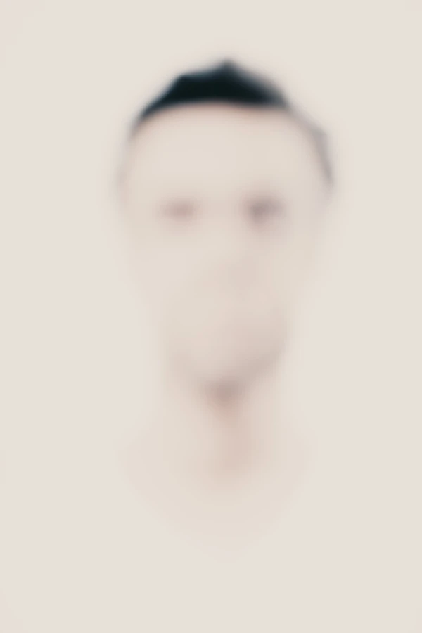 Portrait high-key défocalisé : visage d’homme vu de face, cheveux courts foncés, contours et traits volontairement estompés sur fond crème minimal — série « Erasure ».