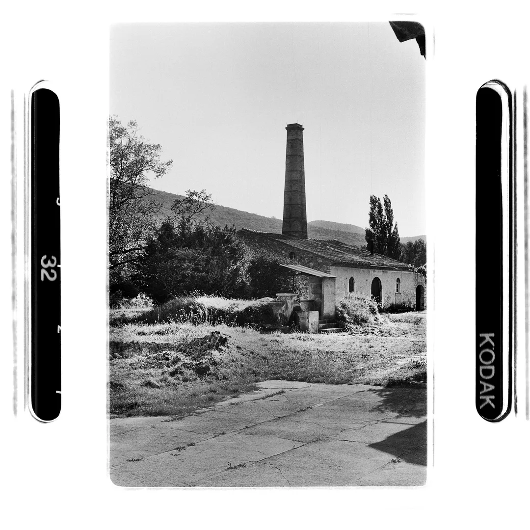 Photo noir et blanc de la grande cheminée industrielle au poët-laval dans la Drôme, devant les anciens bâtiments liés à l’activité de poterie, réalisée dans le cadre du reportage sur le potier Jacky Robin et la terre blanche du village.