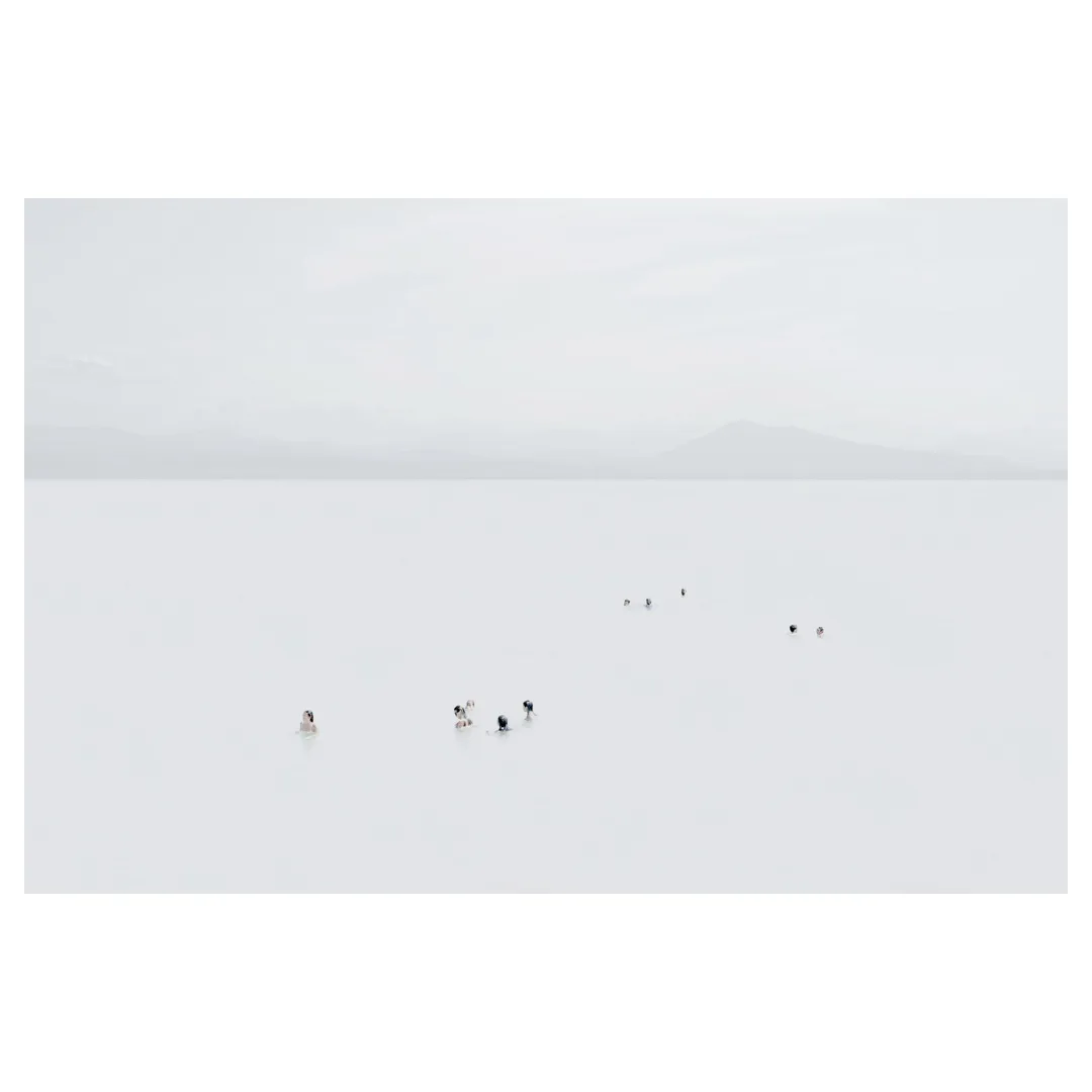 Photographie high-key en plan très large : petits groupes de nageurs alignés dans une mer laiteuse, horizon net et reliefs montagneux diffus au loin, composition minimaliste centrée sur l’espace et la profondeur esthétique.