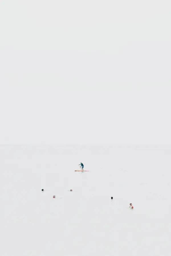 Photographie high-key d’un paysage marin en plan large : pratiquant de stand up paddle isolé sur mer calme, bouées disséminées et quelques baigneurs, horizon effacé, composition minimaliste axée sur l’espace et l’échelle humaine.
