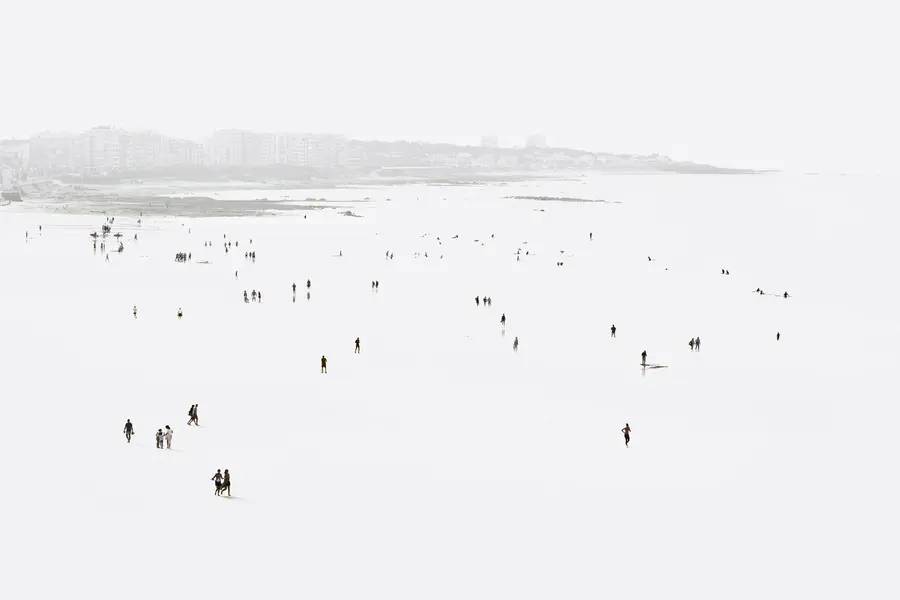 Photographie d’exposition high-key en grand angle d’une plage à marée basse : silhouettes éparses sur sable humide, reflets subtils, horizon urbain brumeux, composition minimaliste jouant sur le vide et l’échelle humaine.
