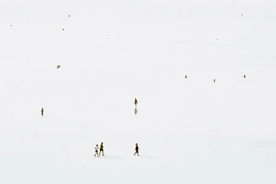 Photographie high-key en plan large : quelques marcheurs et baigneurs disséminés sur une étendue de sable et d’eau très claire, reflets subtils, composition minimaliste centrée sur l’espace négatif et le rythme des figures humaines.