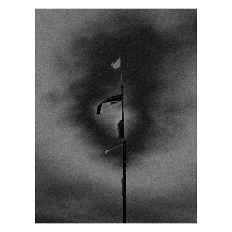 Photographie en noir et blanc d’un mât de plage avec plusieurs drapeaux et bandes de signalisation battus par le vent, cadré en vertical sur un ciel sombre et orageux, série « Marée intérieure » de Nicolas Louis.