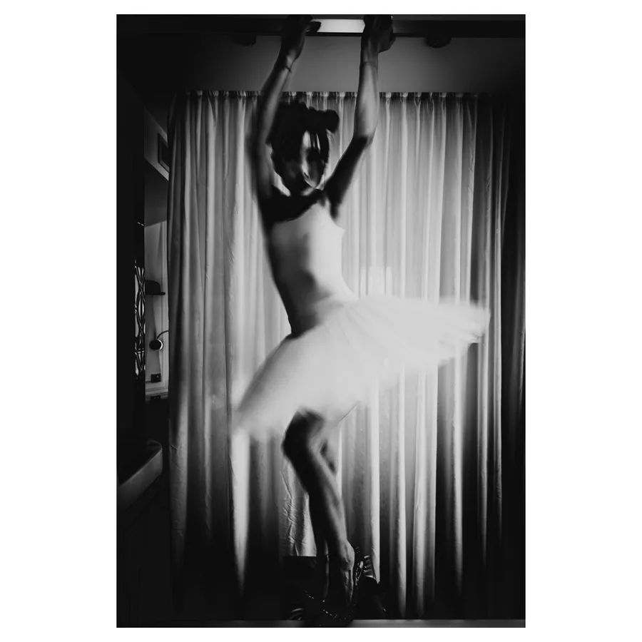 Lala en danse, bras levés accroché à une barre, flou artistique, tutu en rotation devant des rideaux, d’une salle de bain en noir et blanc.