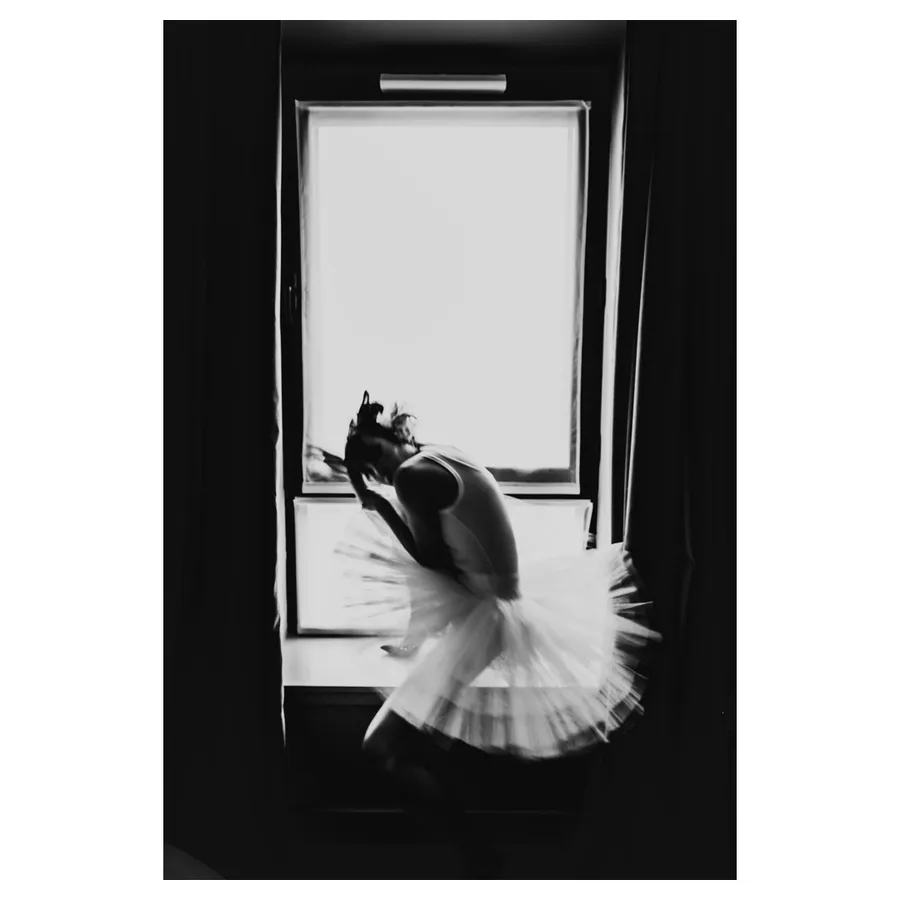 Lala penchée près du cadre de la fenêtre, flou de mouvement, tutu en éventail, regardant le photographe avec un rendu monochrome.
