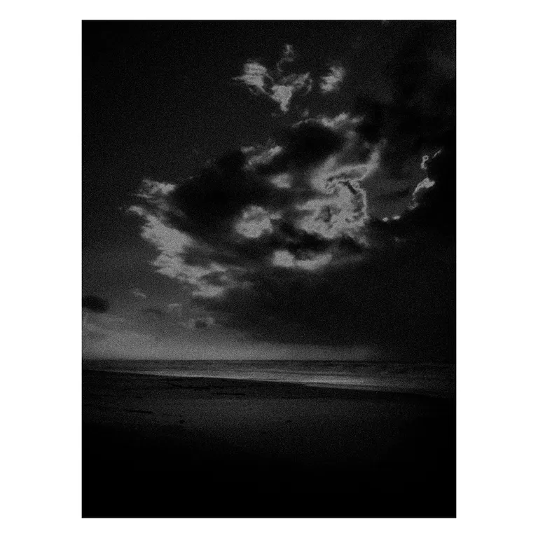 Photographie en noir et blanc très contrastée d’une plage atlantique au crépuscule, vaste ciel sombre avec amas de nuages éclairés par l’arrière, ligne de mer basse et sable obscur au premier plan, série « Marée intérieure » de Nicolas Louis.