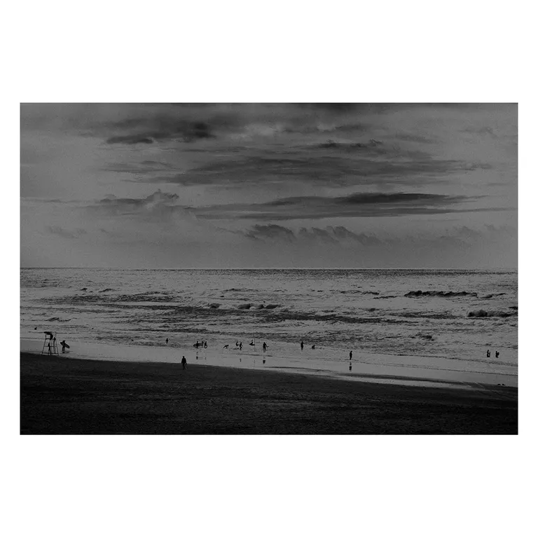 Photographie noir et blanc d’une plage du Porge : ciel chargé, Atlantique houleux, silhouettes de baigneurs dispersées et poste de secours à gauche, série « Marée intérieure ».