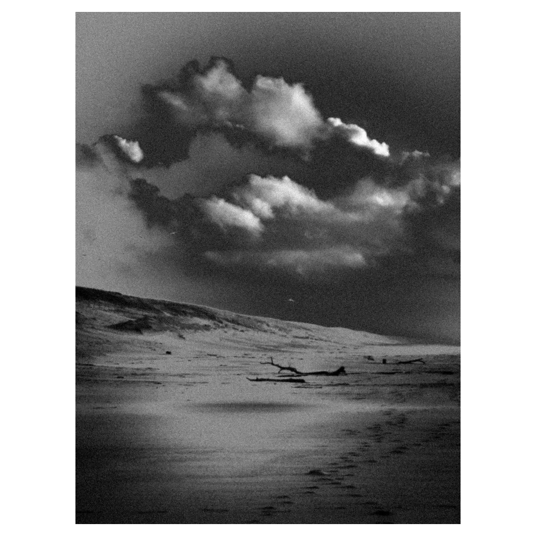 Photographie noir et blanc d’une plage déserte : dunes, ciel orageux au-dessus d’un rivage sableux, bois flotté et traces de pas dans le sable dans un effet de rendu très esthétique pour une exposition photo.