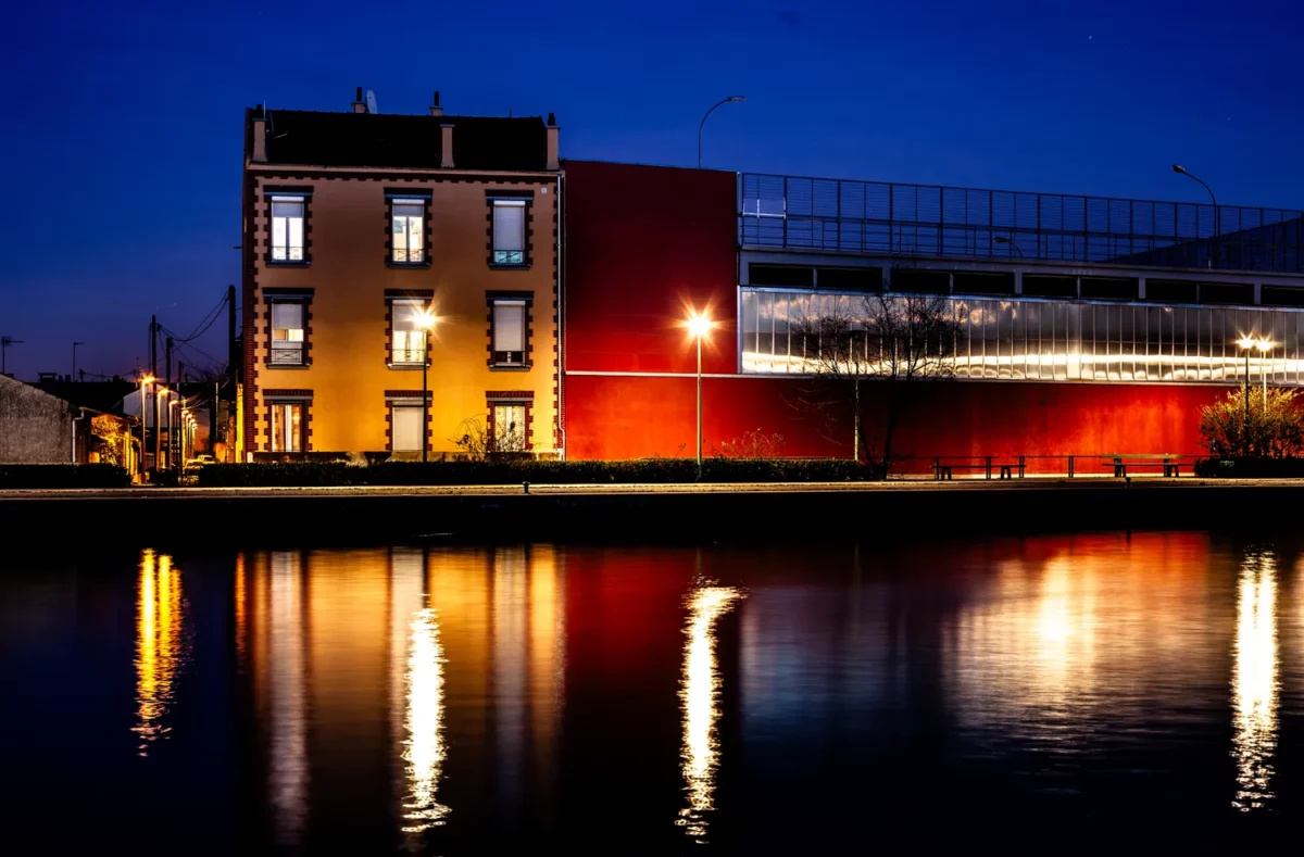 Photographie d’architecture nocturne sur les bords du canal de Saint Denis, avec façade ancienne et bâtiment contemporain rouge se reflétant dans l’eau, réalisée par un photographe professionnel à Paris