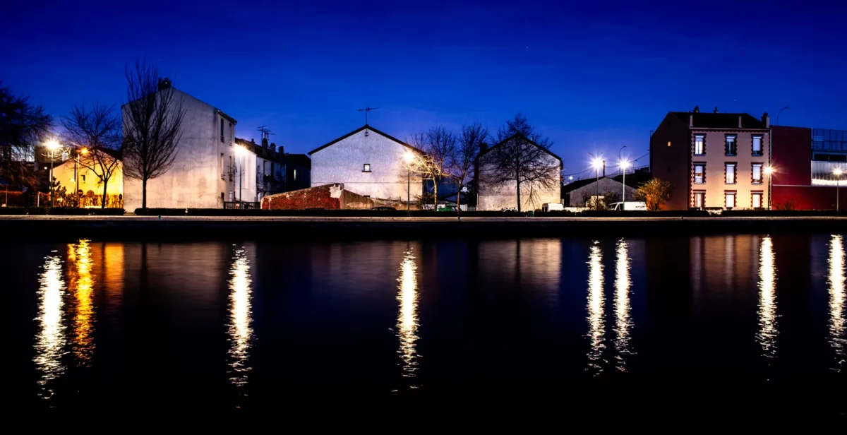 Photographie nocturne d’architecture en bord du canal Saint Denis, avec rangée de maisons éclairées et reflets des lampadaires dans l’eau, réalisée par un photographe professionnel à Paris