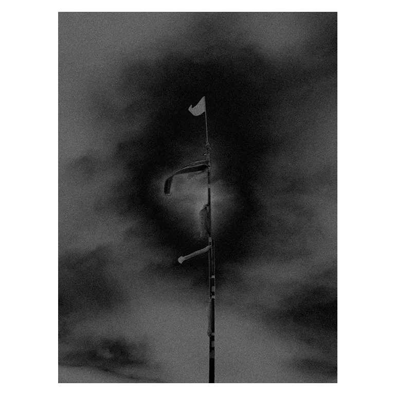 Photographie en noir et blanc d’un mât de plage du Porge, avec plusieurs drapeaux et bandes de signalisation battus par le vent, cadré en vertical sur un ciel sombre et orageux, série « Marée intérieure » de Nicolas Louis.