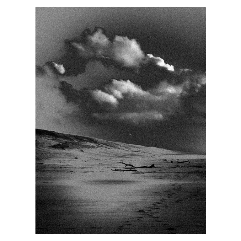 Photographie noir et blanc d’une plage déserte, au porge : dunes, ciel orageux au-dessus d’un rivage sableux, bois flotté et traces de pas dans le sable dans un effet de rendu très esthétique pour une exposition photo.