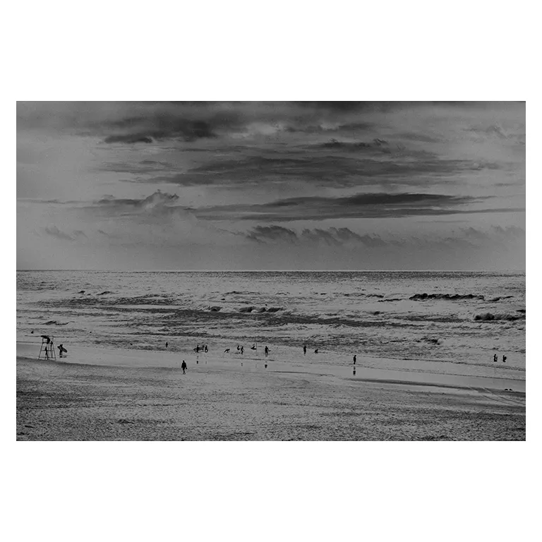 Photographie noir et blanc d’une plage du Porge : ciel chargé, Atlantique houleux, silhouettes de baigneurs dispersées et poste de secours à gauche, série « Marée intérieure ».