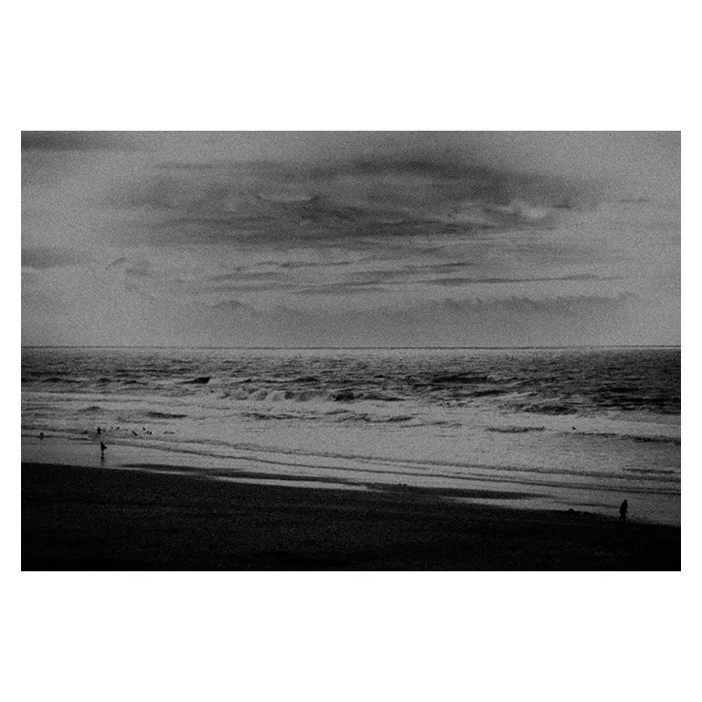 Photographie noir et blanc très contrastée d’une plage en Gironde : plaques d’écume au premier plan sur le sable sombre, miroir d’eau calme vers l’horizon, vagues et nuages orageux en arrière-plan, série « Marée intérieure » de Nicolas Louis.