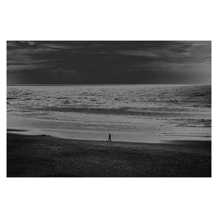 Photographie en noir et blanc d’une plage atlantique au ciel très sombre, mer animée par de petites vagues, une silhouette seule marche sur le sable au centre inférieur du cadre, série « Marée intérieure » de Nicolas Louis.