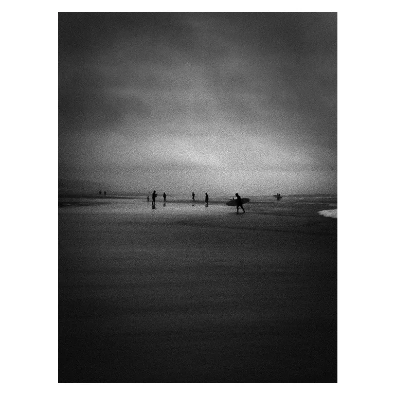 Photographie en noir et blanc de l’Atlantique à marée basse : silhouettes de surfeurs et de baigneurs dispersées dans l’eau peu profonde, un surfeur portant sa planche au premier plan, atmosphère brumeuse et tonalités très contrastées, série « Marée intérieure » de Nicolas Louis.