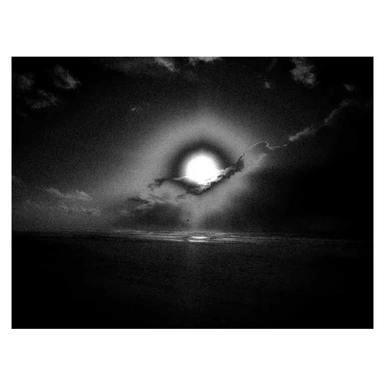 Soleil bas avec un halo circulaire perçant les nuages au-dessus de l’océan à Porge plage pour une photographie en noir et blanc très contrastée destinée à une exposition photo.
