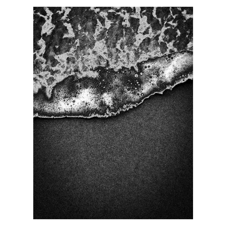 Gros plan en noir et blanc d’une vague qui vient mourir sur le sable sombre de Porge plage, écume détaillée en bordure formant une ligne diagonale, photographie graphique de la série « Marée intérieure » de Nicolas Louis.