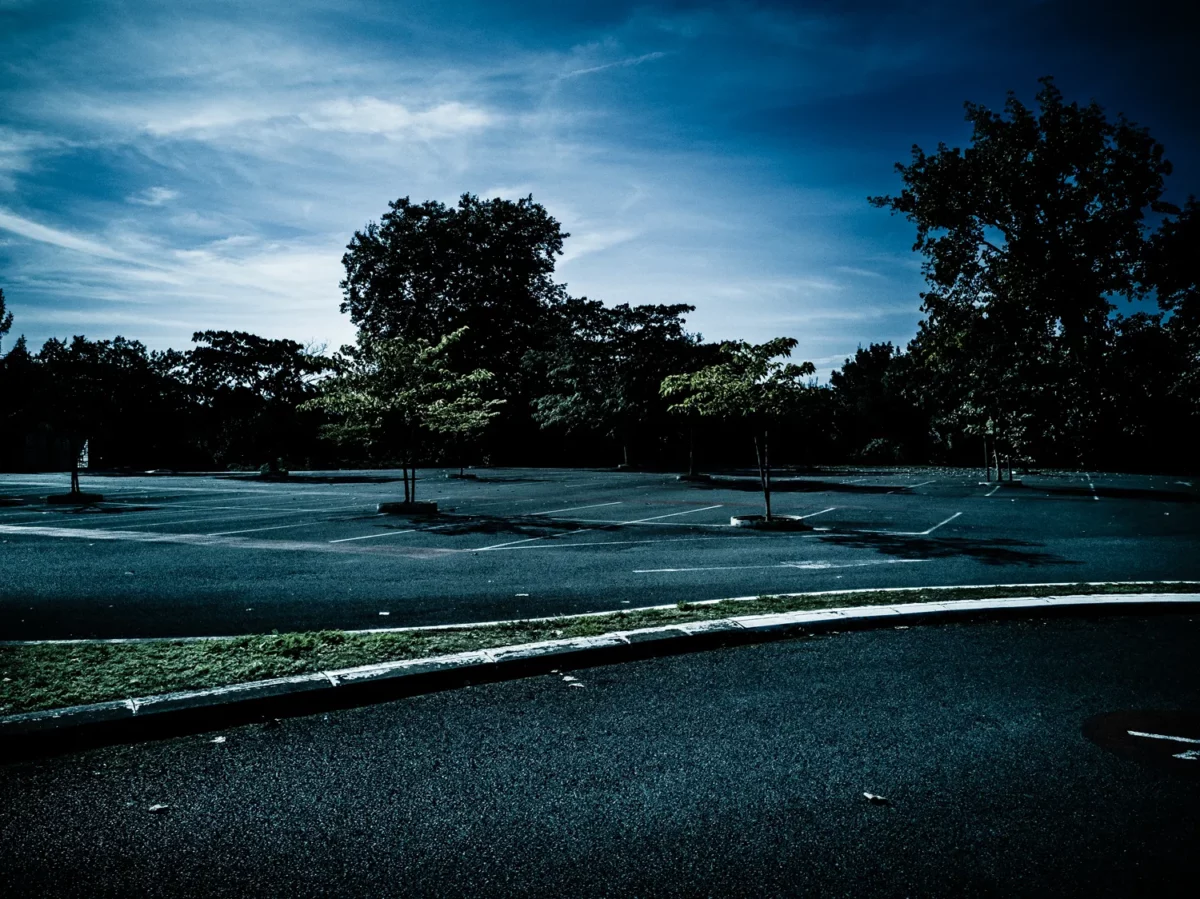 Photographie de paysage urbain d’un grand parking vide bordé d’arbres sous ciel bleu, réalisée par un photographe artiste.