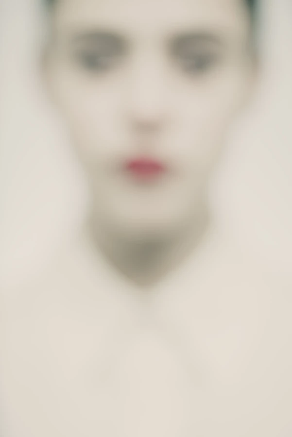 Portrait artistique high-key de Louise, net volontairement absent : gros plan frontal d’un visage féminin, traits et regard floutés, lèvres rouge vif au centre, chemise claire fermée — série « Erasure ».