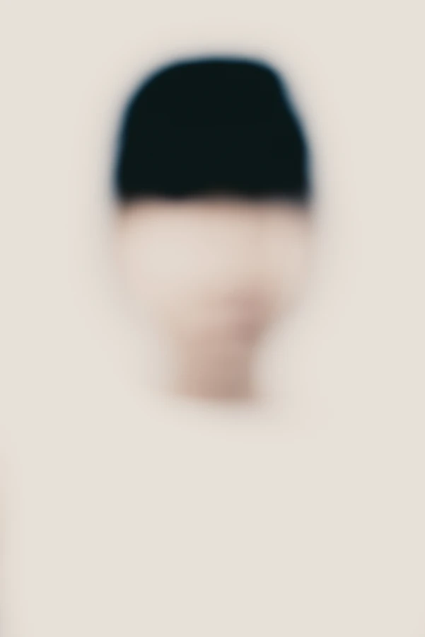 Portrait artistique high-key, net volontairement absent : visage de face très flou, masse noire géométrique en haut du cadre (coiffe ou chevelure), fond crème minimaliste — série « Erasure ».