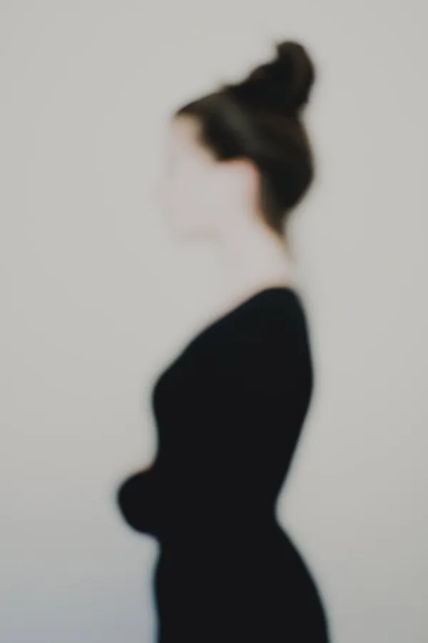 Photographie artistique défocalisée : silhouette féminine de profil, chignon et tenue sombre, contours adoucis et identité estompée, fond neutre minimaliste — extrait de la série « Erasure »