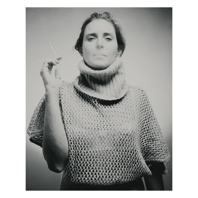 Photographie plasticienne en studio présentant un portrait de femme en pull à col roulé, tenant une cigarette et regardant fixement le photographe, avec un traitement noir et blanc intemporel, exploration des volumes et de la lumière naturelle.