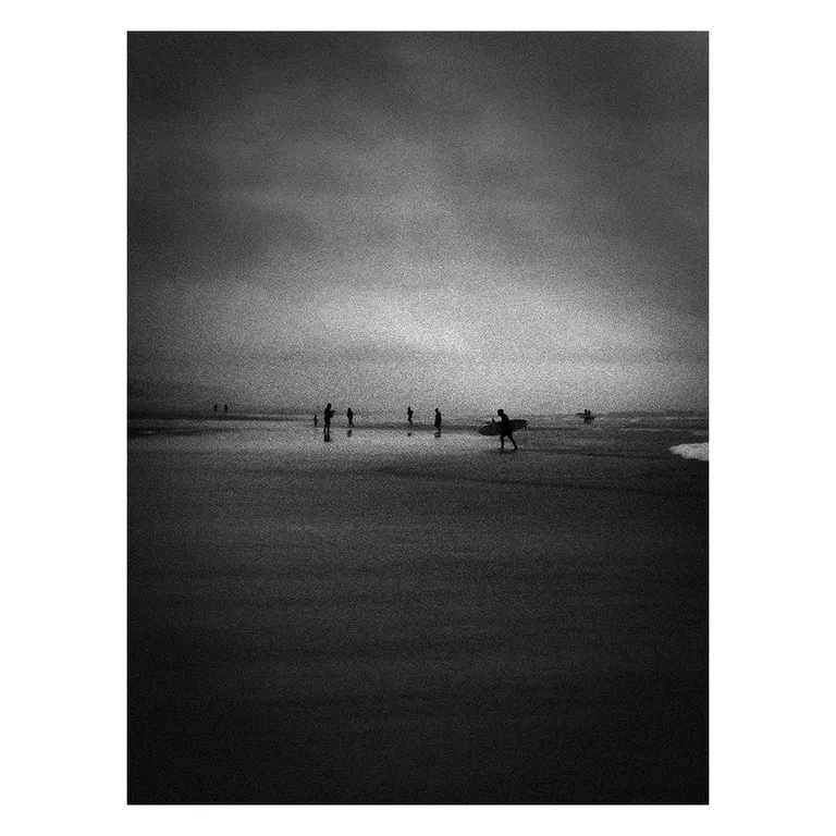 Photographie en noir et blanc de l’Atlantique à marée basse : silhouettes de surfeurs et de baigneurs dispersées dans l’eau peu profonde, un surfeur portant sa planche au premier plan, atmosphère brumeuse et tonalités très contrastées, série « Marée intérieure » de Nicolas Louis.