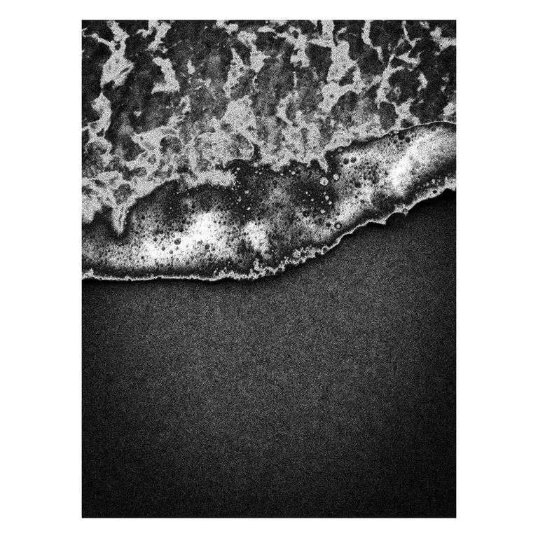 Gros plan en noir et blanc d’une vague qui vient mourir sur le sable sombre, écume détaillée en bordure formant une ligne diagonale, photographie graphique de la série « Marée intérieure » de Nicolas Louis.
