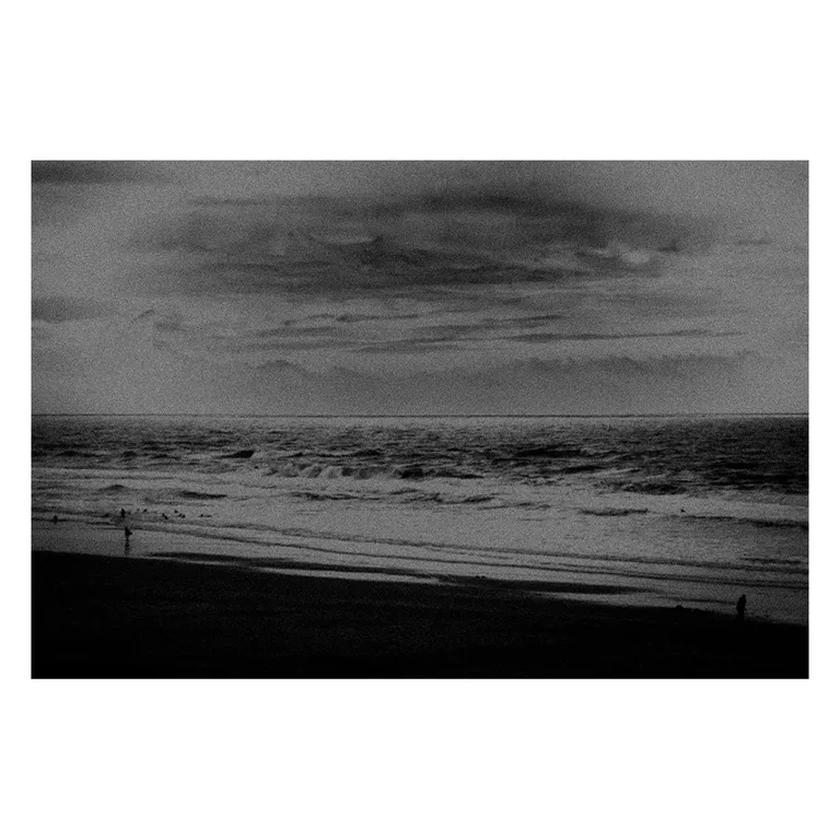 Photographie en noir et blanc d’une plage atlantique vue de haut, mer agitée avec plusieurs lignes de vagues, ciel nuageux strié, quelques silhouettes de promeneurs en bas de cadre, série « Marée intérieure » de Nicolas Louis.