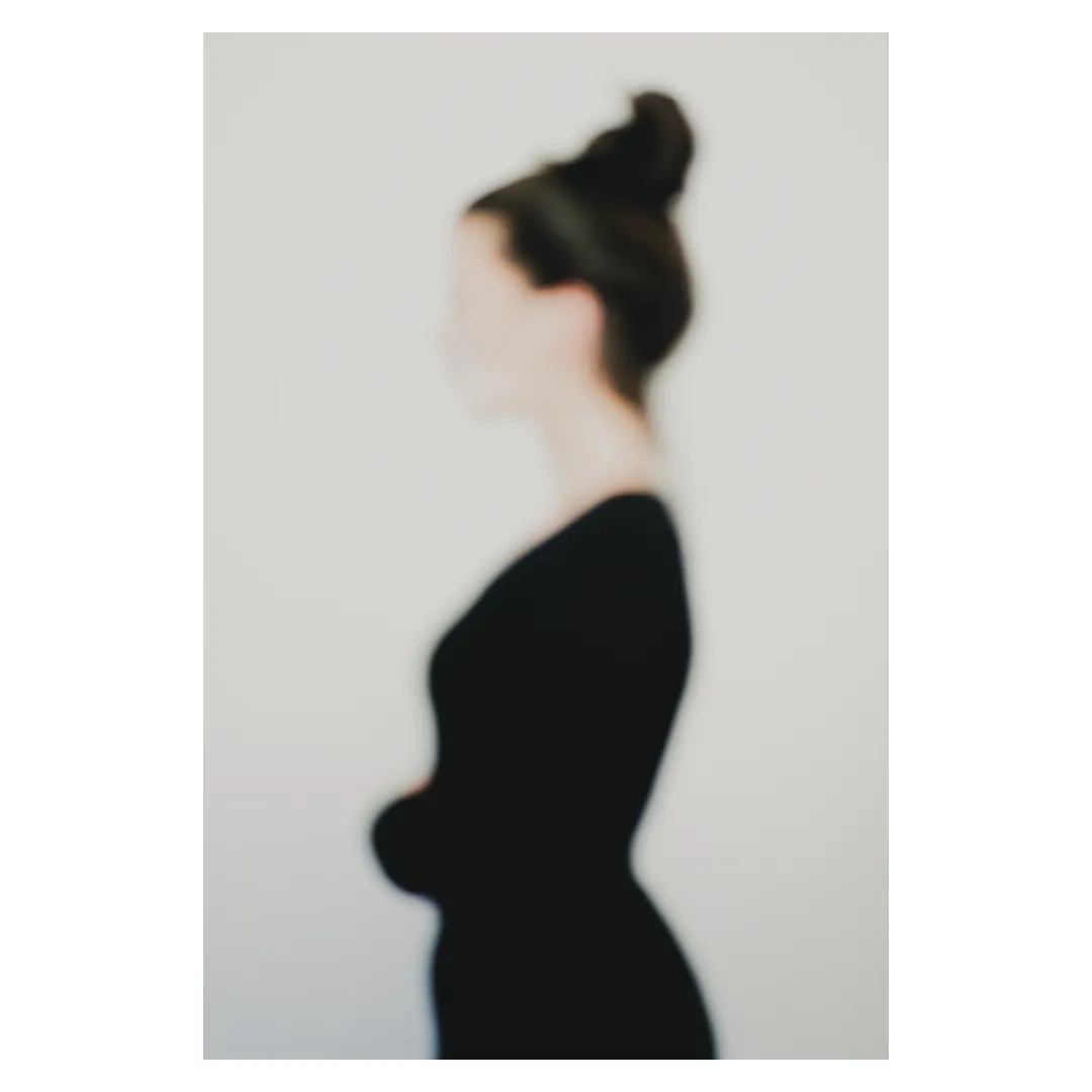 Photographie artistique défocalisée : silhouette féminine de profil, chignon et tenue sombre, contours adoucis et identité estompée, fond neutre minimaliste — extrait de la série « Erasure »