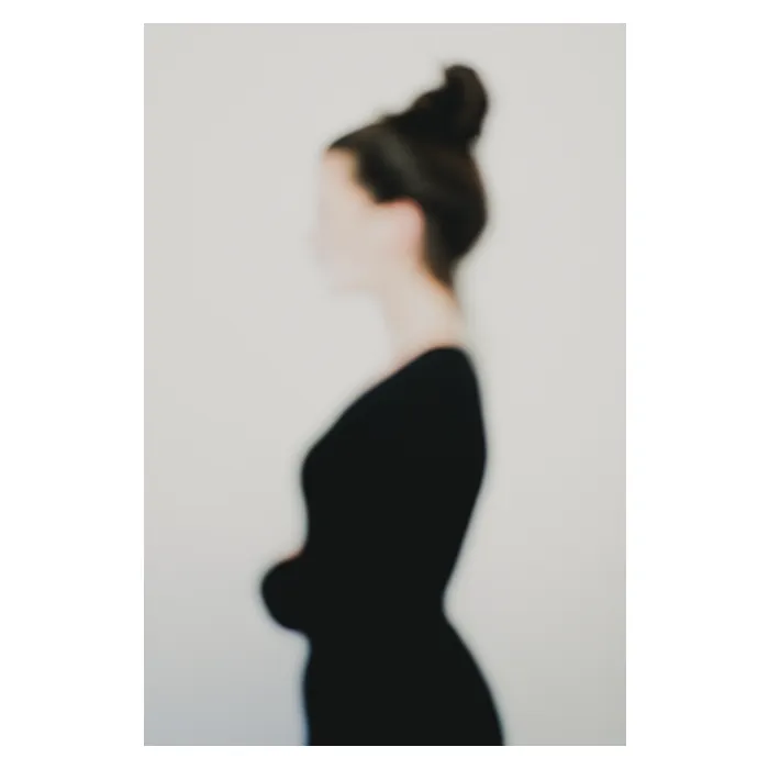 Photographie artistique défocalisée : silhouette féminine de profil, chignon et tenue sombre, contours adoucis et identité estompée, fond neutre minimaliste — extrait de la série de portrait « Erasure », par le photographe Nicolas Louis
