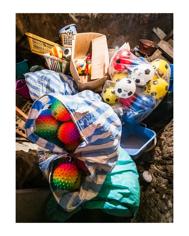 Vue plongeante sur un bric-à-brac coloré : ballons arc-en-ciel dans un sac rayé bleu et blanc, ballons de football jaunes et rouges, paniers plastique orange, cartons et tissus entassés aux Grands Magasins Universels de Dieulefit