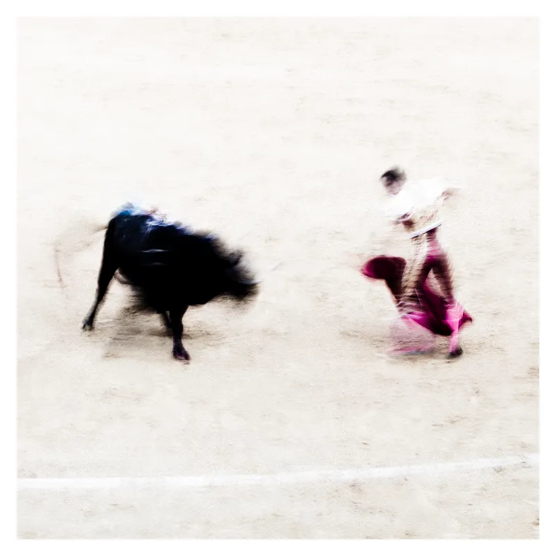 Photographie floue artistique d'une corrida à Bayonne montrant un taureau noir et un torero en costume rose fuschia dans l'arène, effet de mouvement pictural sur fond beige clair