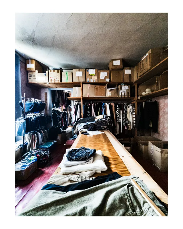 Dressing spacieux aux Grands Magasins Universelles de Dieulefit : vêtements suspendus sur portants muraux, table centrale en bois avec piles de vêtements soigneusement pliés, cartons de rangement étiquetés au plafond, lumière naturelle douce créant une ambiance chaleureuse