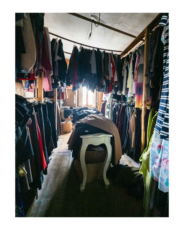 Intérieur d'un dressing encombré sous verrière aux Grands Magasins Universels de Dieulefit, Drôme. Vêtements colorés suspendus de part et d'autre, lumière naturelle diffuse créant un contraste dramatique avec les ombres profondes, chaise ancienne au centre