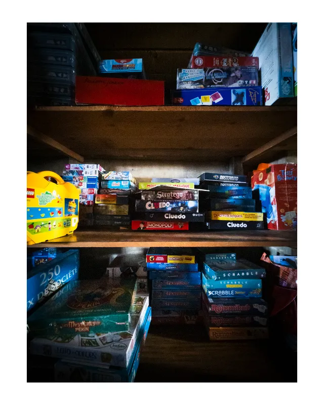 Trois étagères en bois sombre exposant une collection de jeux de société classiques - Monopoly, Cluedo, Scrabble, Stratego - captées en lumière naturelle douce créant une ambiance nostalgique dans un magasin traditionnel de Dieulefit
