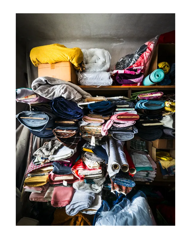 Étagères en bois chargées de tissus et textiles multicolores soigneusement pliés et empilés, capturées en lumière naturelle dans l'atmosphère authentique des Grands Magasins Universelles à Dieulefit