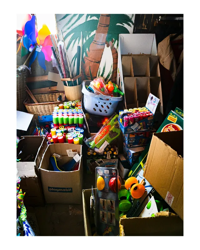 Composition photographique d'un bric-à-brac artistique aux grands magasins universels de Dieulefit : tubes de peinture multicolores, pinceaux dans un pot en céramique, panneaux tropicaux, jouets Playmobil et fournitures créatives disposés dans des cartons et paniers en osier, éclairage naturel contrasté