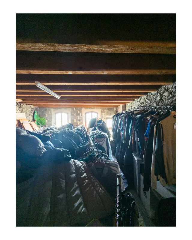Intérieur chaleureux d'une boutique de vêtements à Dieulefit dans la Drôme, avec portants chargés de vestes et manteaux bleus, plafond à poutres en bois apparentes, murs en pierre et lumière naturelle diffuse traversant les fenêtres
