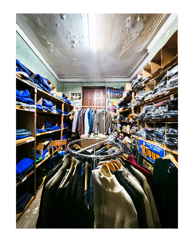 Vue grand-angle de l'intérieur d'une boutique de vêtements traditionnelle à Dieulefit dans la Drôme, avec étagères en bois chargées de textiles bleus et naturels, plafond ancien patiné, portant central de pulls et vestes colorées