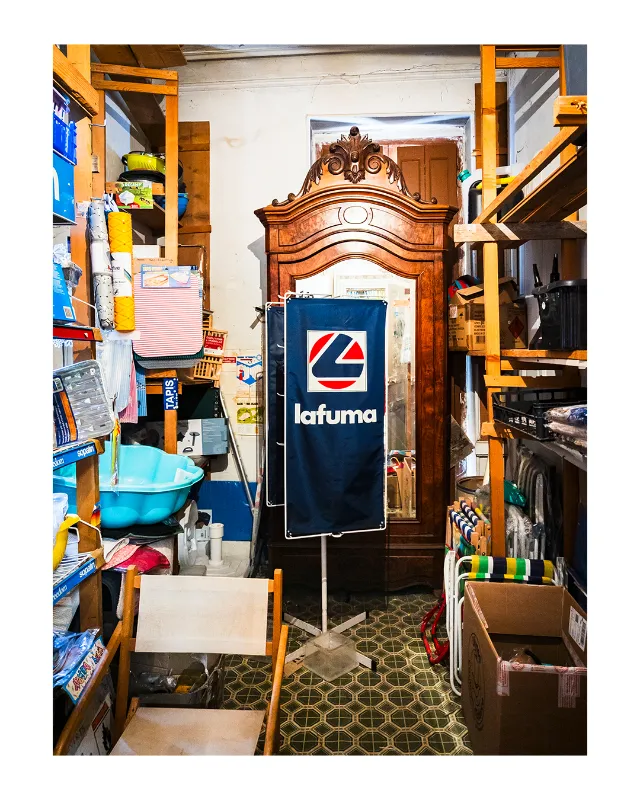 Photographie d'intérieur du magasin vintage Les Grands Magasins Universels à Dieulefit dans la Drôme, armoire en bois sculpté ornementale au centre, drapeau Lafuma bleu au premier plan, étagères colorées chargées de produits, sol carreaux géométriques verts